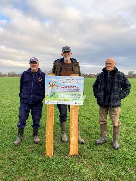 Colin Ballard (Upton Town Council), John Dickinson (Worcester Curlew Group) amd Mike Smart (Curlew Action) erecting dog-friendly signs on Upton Ham, February 2022. (Photo by John Dickinson) A photo of three men standing in a field with a sign about dogs and ground-nesting birds.