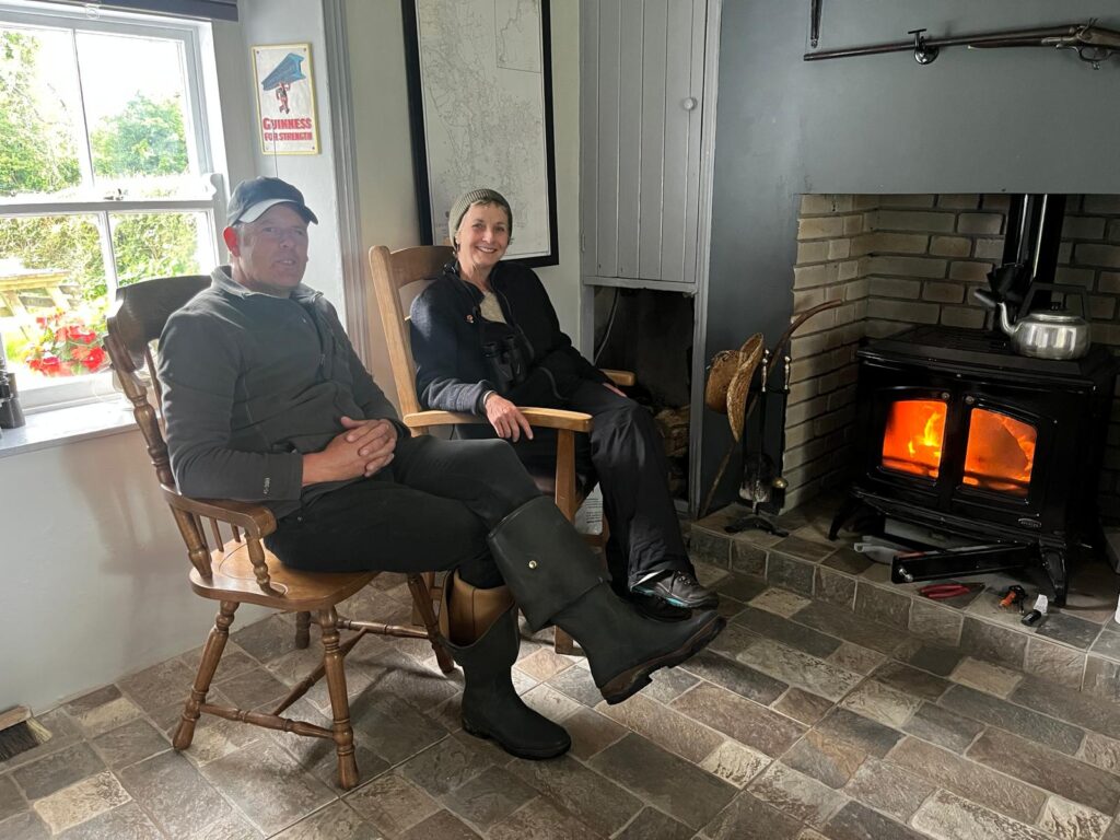 A photo of a man and a woman sat inside, in wooden chairs by a wood burner.
