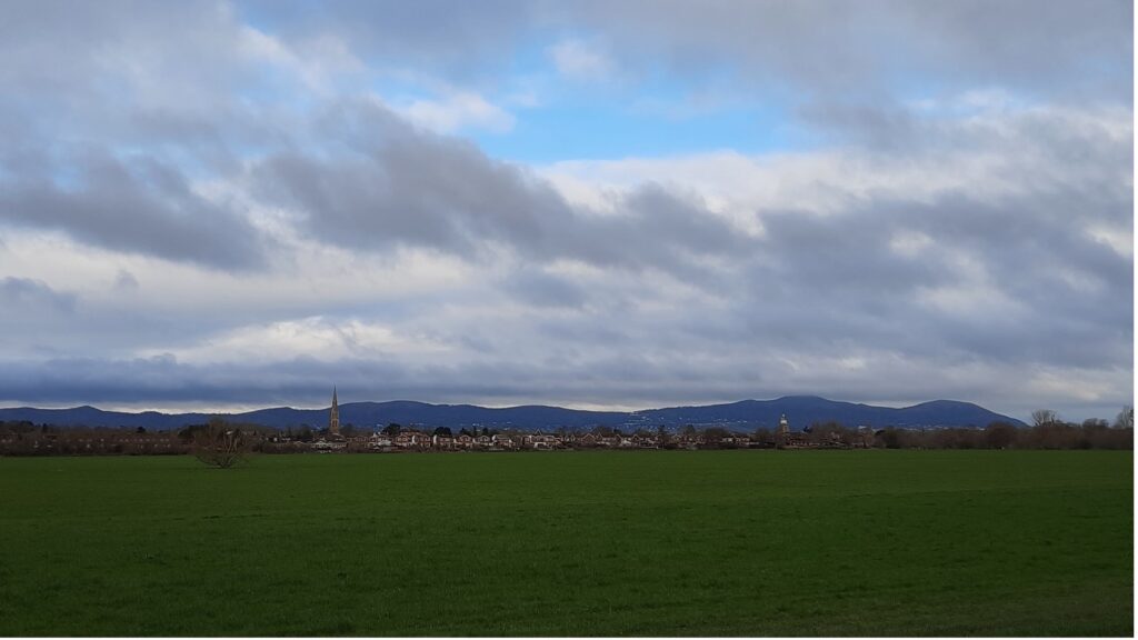 Upton: Ham in the foreground, the town, Malvern Hills in the background. (Photo by John Dickinson) A photo of a field, with a town and hills in the background, and a cloudy sky.